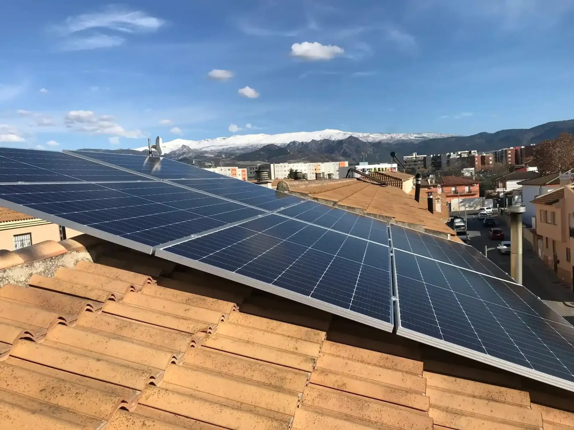 Paneles solares en una azotea bajo un cielo azul con nubes, con vistas a un pueblo y colinas.