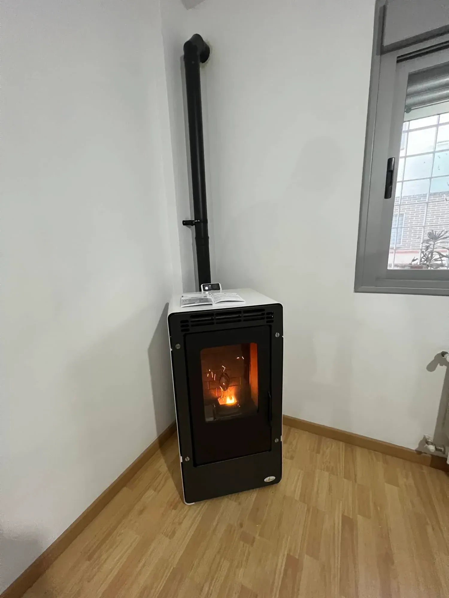Estufa de pellets negra con fuego incandescente en un rincón de una habitación con suelo de madera y paredes blancas.