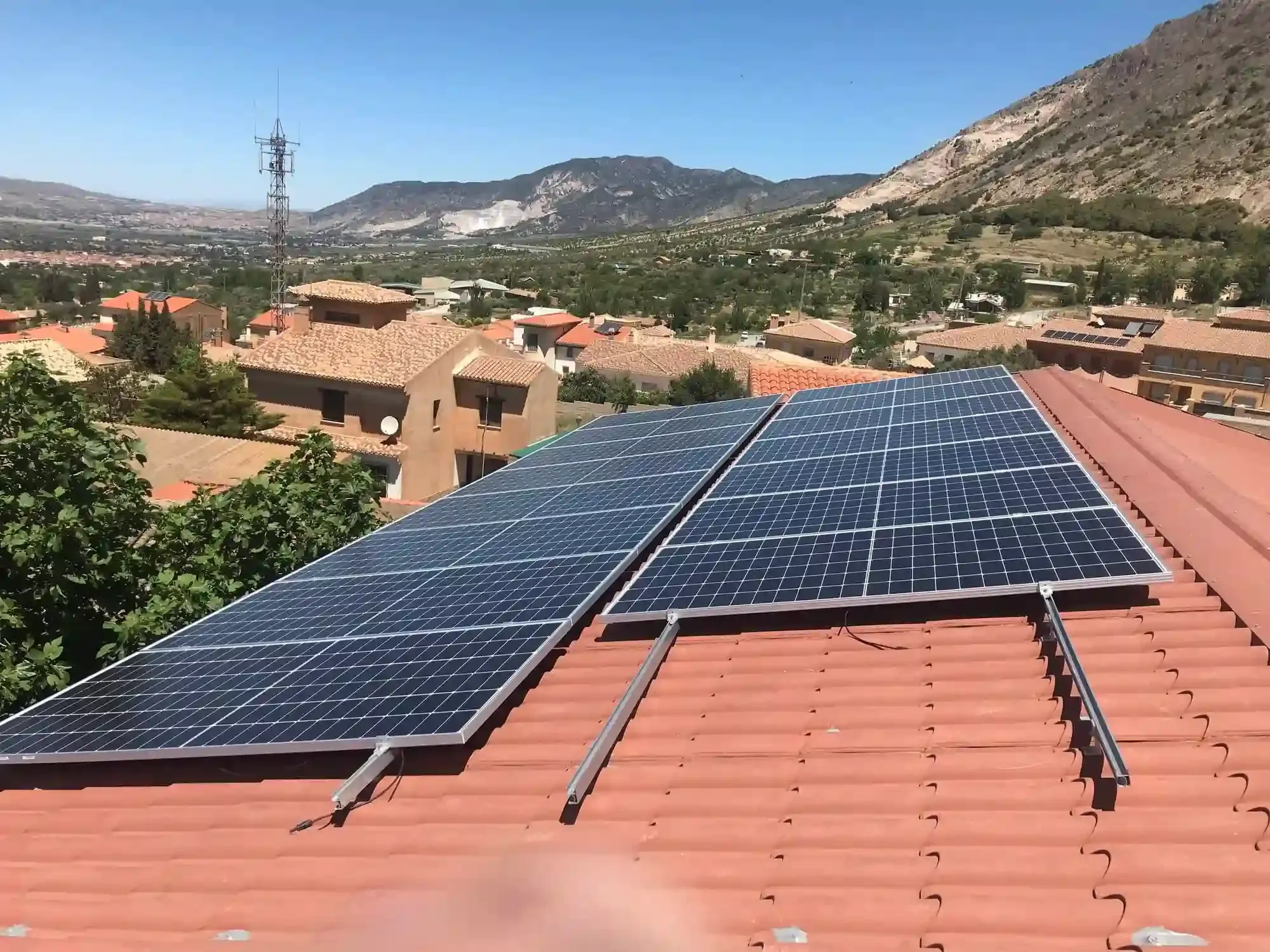 Paneles solares en un tejado de tejas rojas con vistas a un pueblo de montaña bajo un cielo azul despejado.