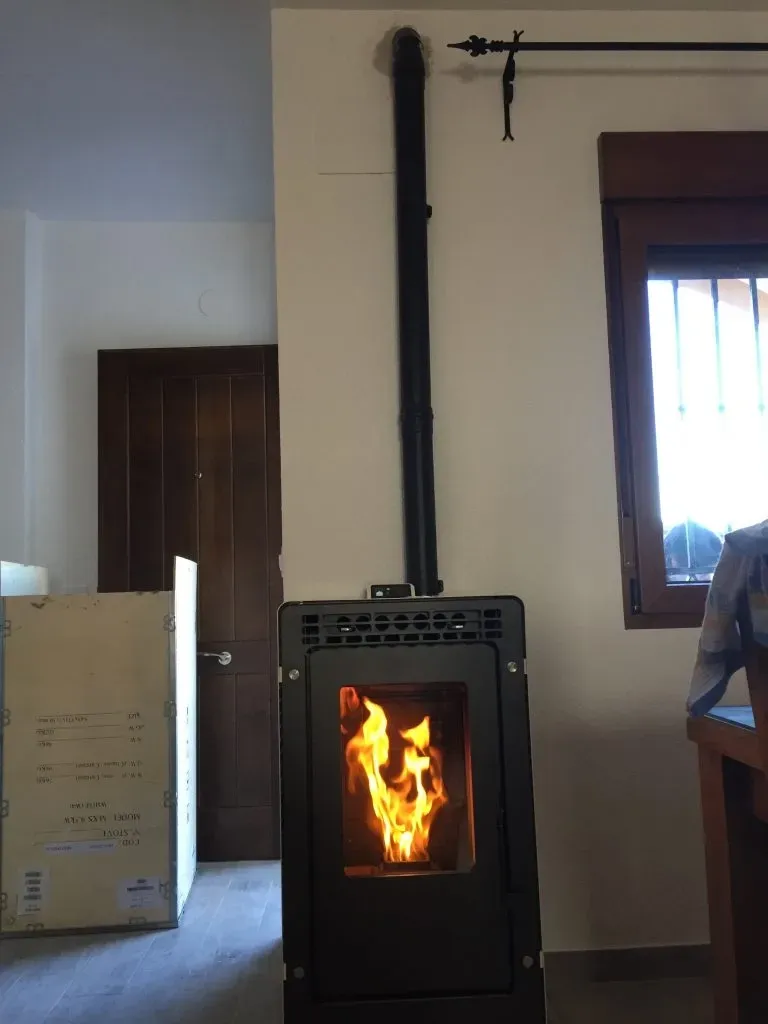 Estufa de leña negra con llama visible en una habitación junto a una ventana y una puerta.