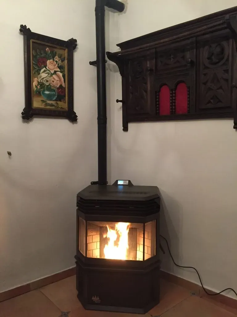 Estufa de leña encendida en un rincón con un estante decorativo en la pared y un cuadro enmarcado sobre un suelo de baldosas.