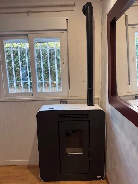 Estufa de leña negra con chimenea en una pequeña habitación junto a una ventana y un espejo de pared.