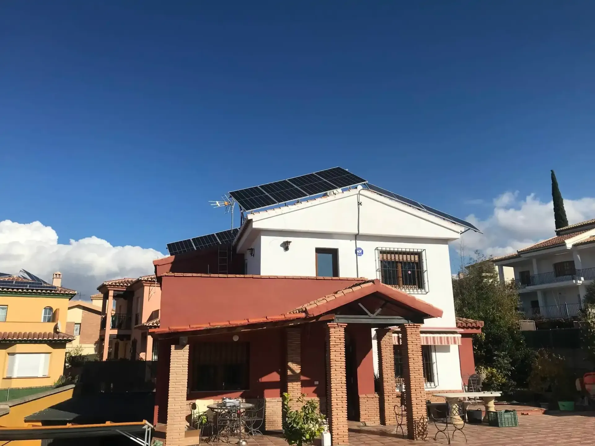 Casa de dos plantas con paneles solares en el tejado y un toldo rojo bajo un cielo azul despejado.