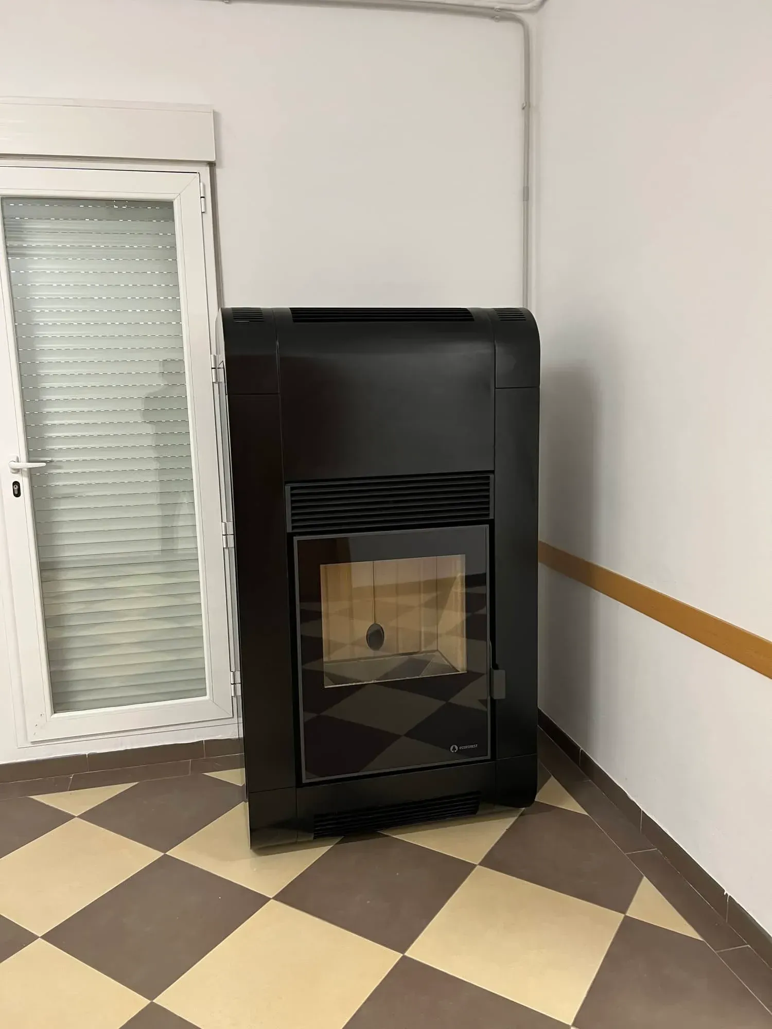 Estufa con frontal de cristal negro en una habitación de esquina con azulejos, junto a una ventana y una pared blanca.