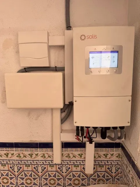 Inversor solar mural con pantalla y cajas eléctricas sobre suelo de baldosas estampadas.
