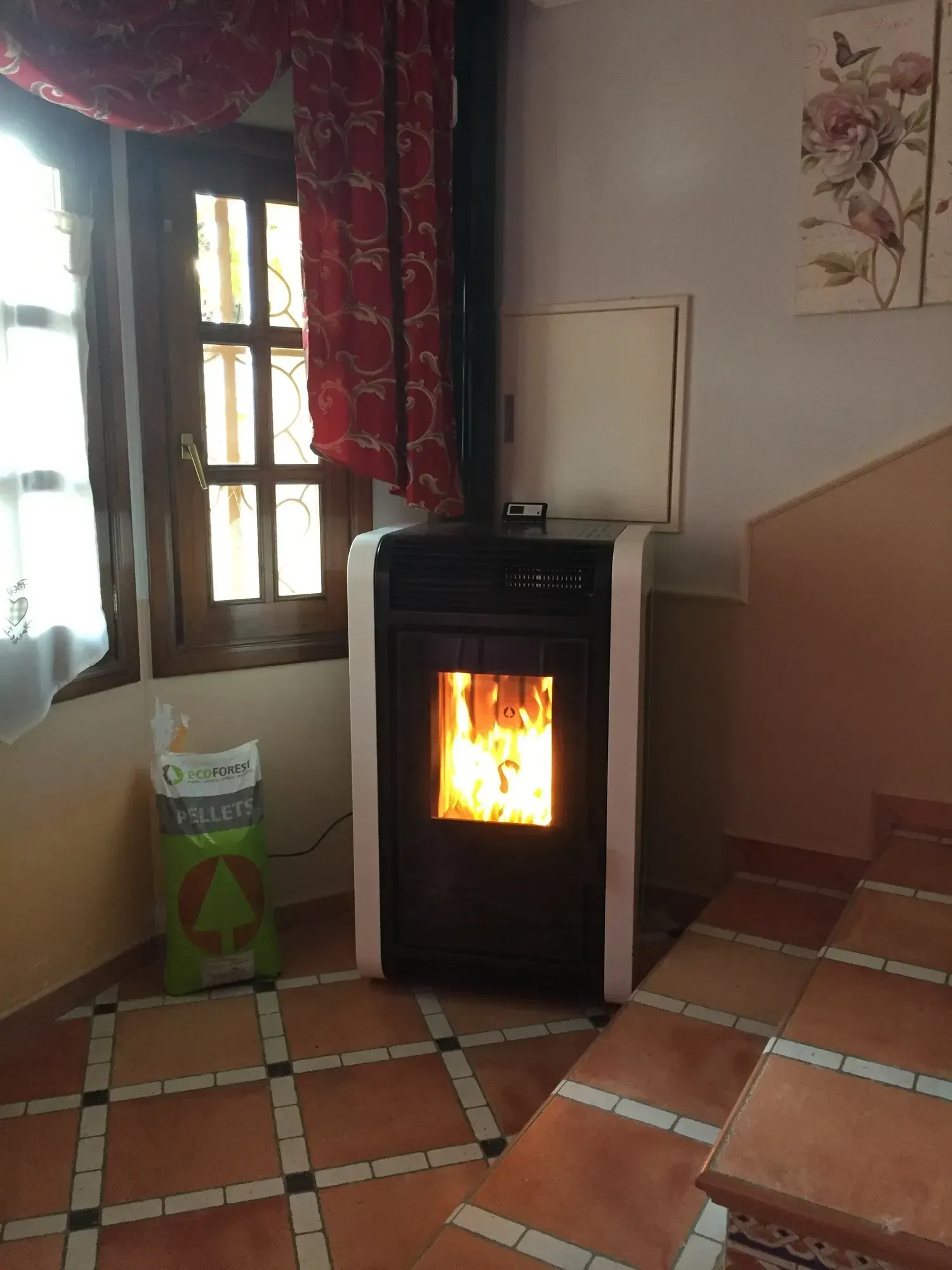 Sala de estar con estufa de pellets negra encendida, suelo de baldosas, ventana y escalera junto a un cuadro floral.