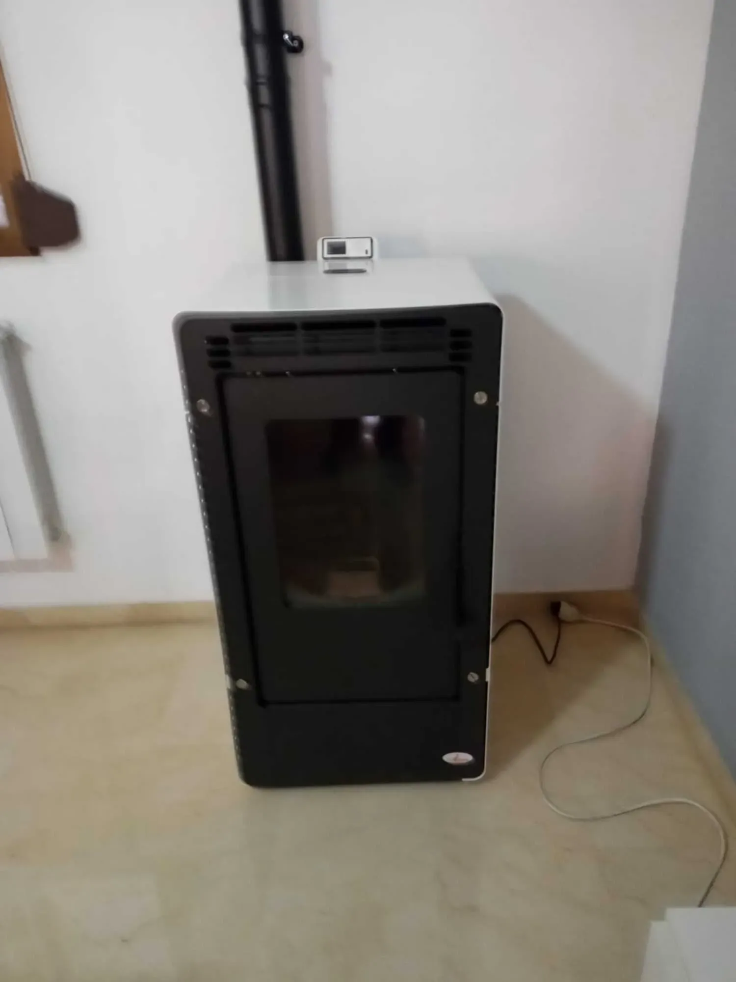 Estufa de pellets de madera negra contra la pared sobre un suelo de baldosas, con un cable de alimentación cerca.