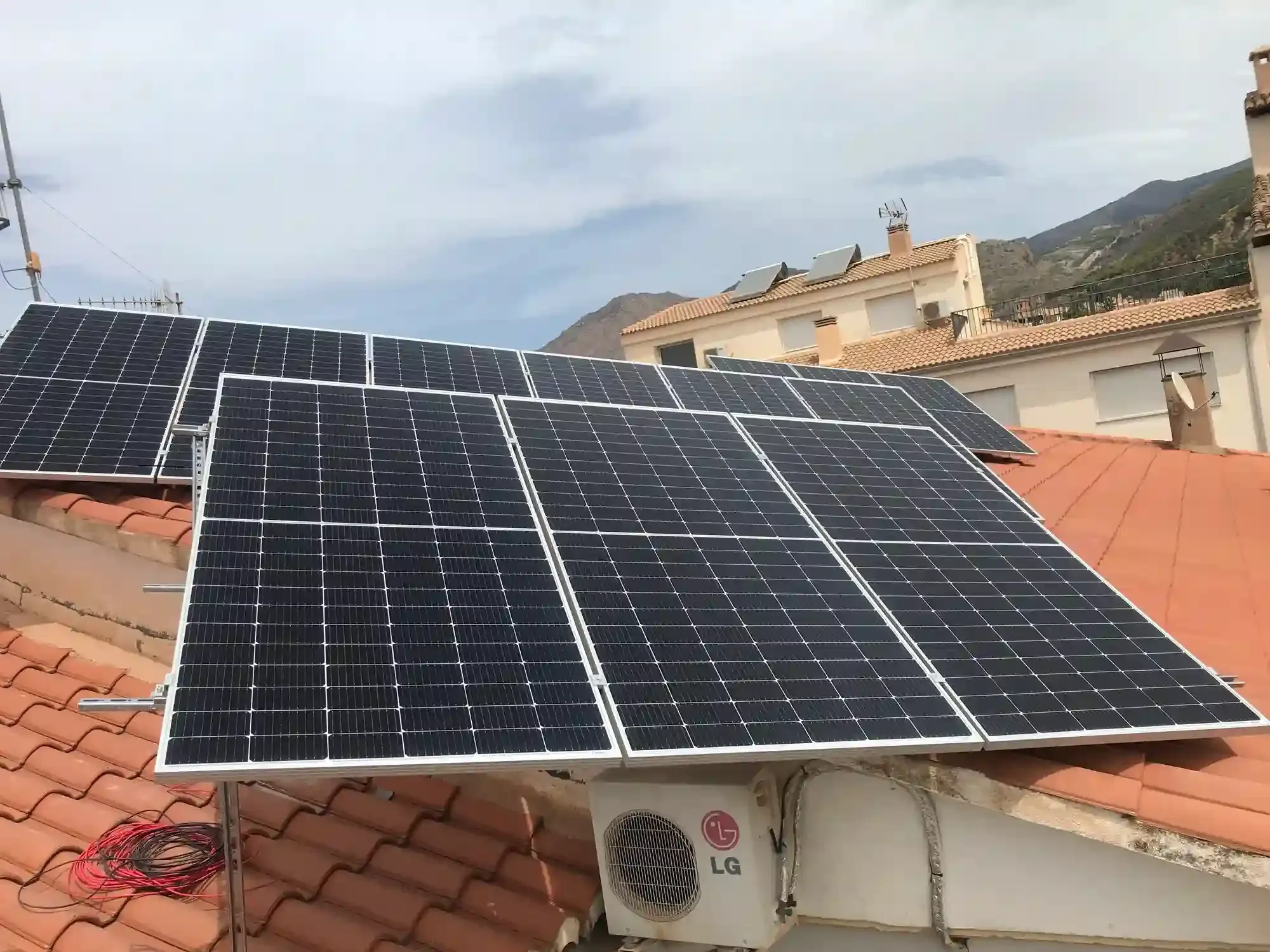 Paneles solares instalados en un tejado de tejas rojas con montañas y edificios al fondo.
