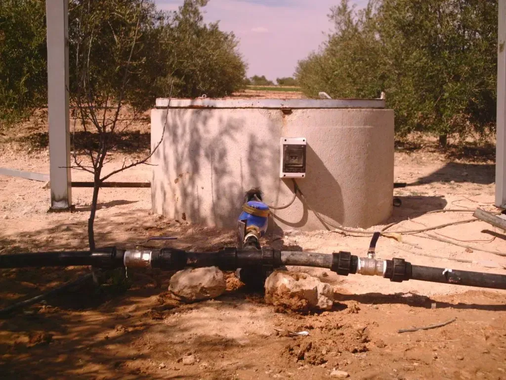 Pequeño depósito de agua y tuberías en una zona seca y arenosa con árboles al fondo.