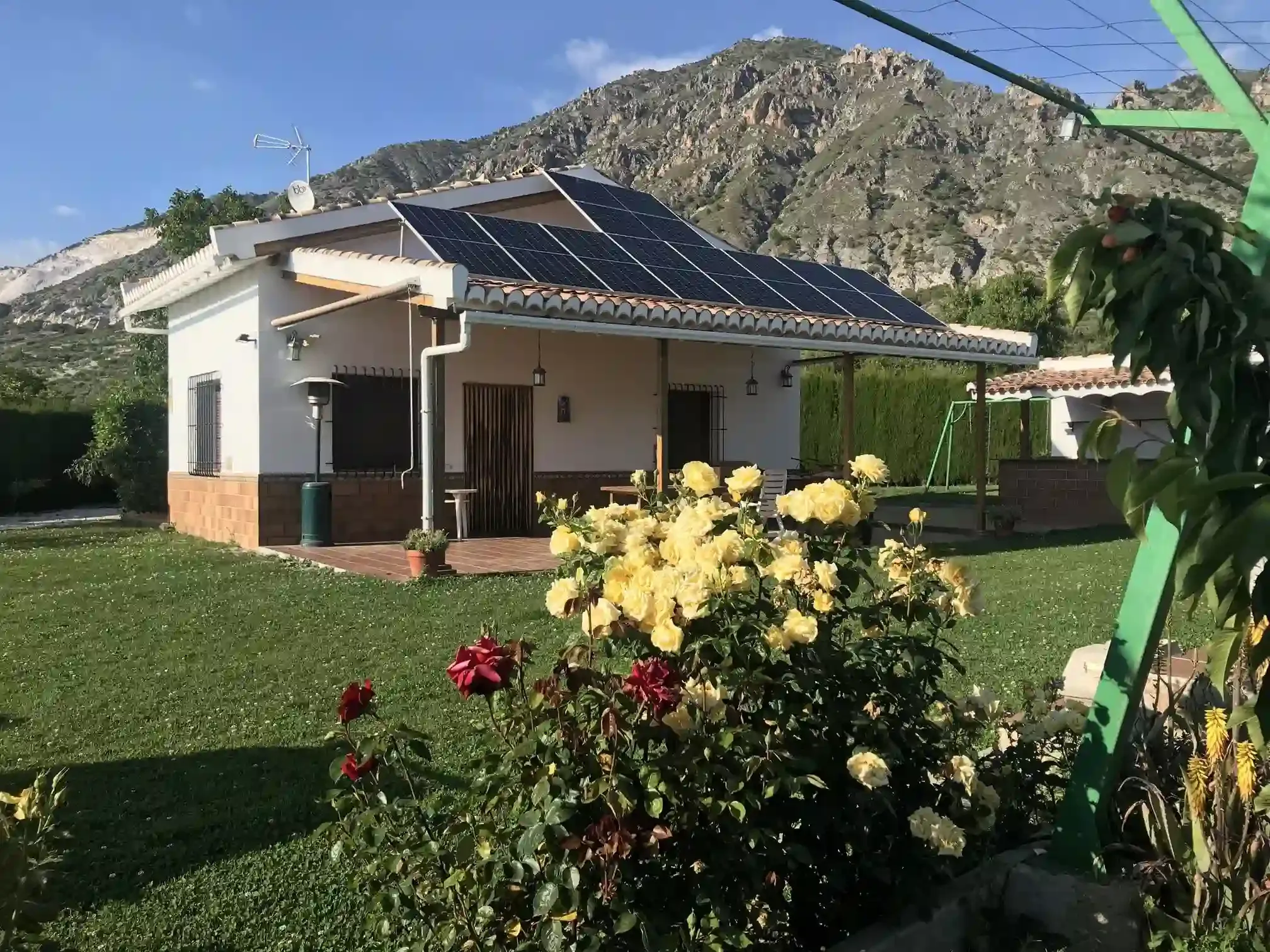 Pequeña casa con paneles solares, rodeada de un césped verde y flores, con montañas al fondo.