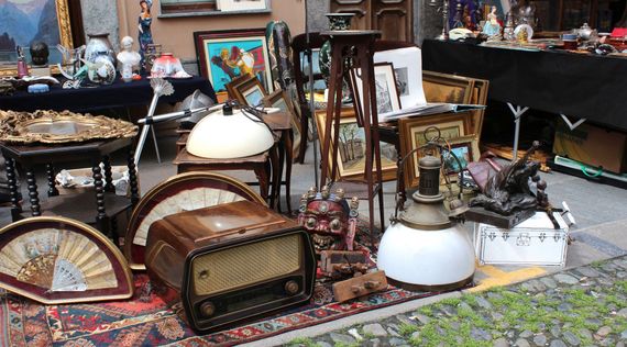 Des radios anciennes, des ventilateurs et des tableaux encadrés sont posés au sol sur un tapis, aux puces en plein air.