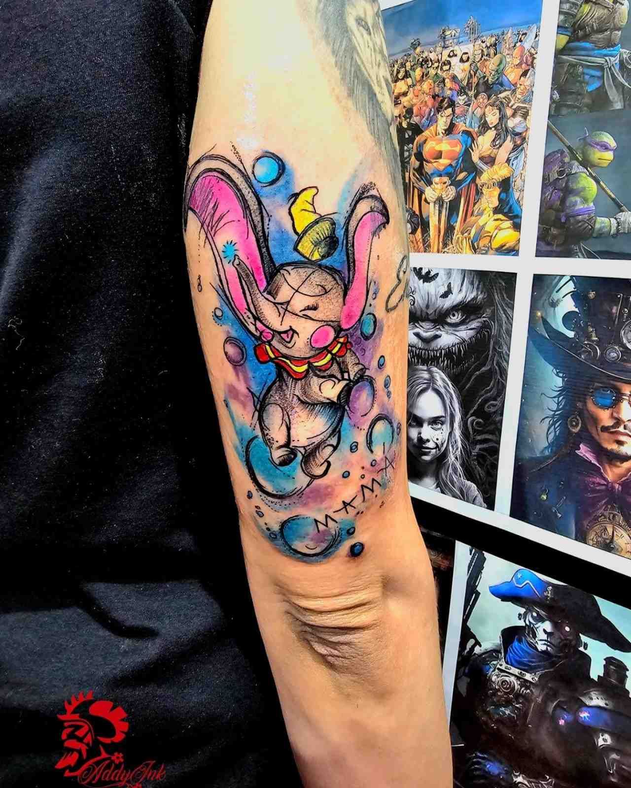 Un tatuaje estilo acuarela de Dumbo, el elefante, en un brazo, con salpicaduras de color azul brillante, rosa y amarillo.