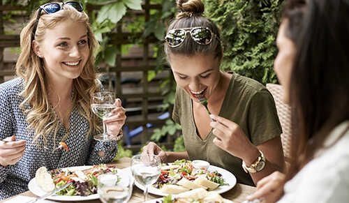 Tres amigos compartiendo una comida y unas bebidas en la mesa de una cafetería al aire libre, sonriendo y charlando