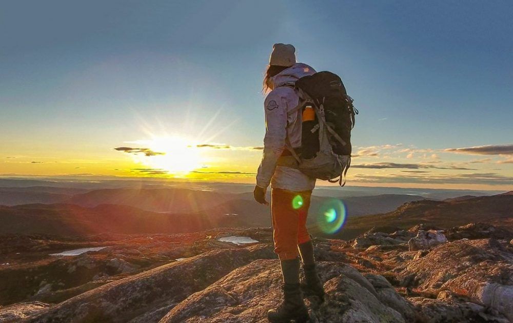 Eine Person mit einem Rucksack steht bei Sonnenuntergang auf einem Berggipfel.