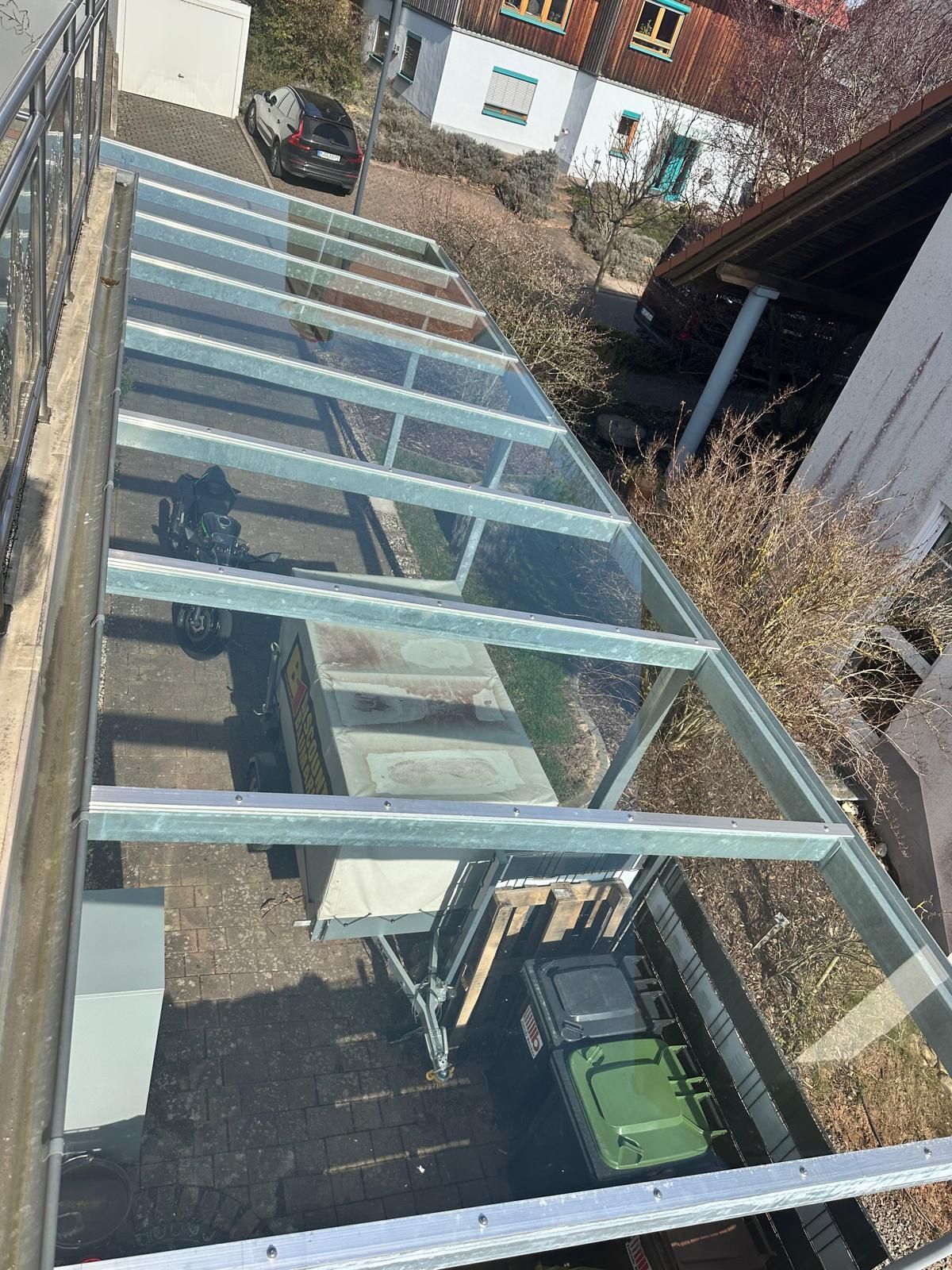Neben einem Haus wird eine verglaste Terrassenüberdachung errichtet, darunter befindet sich eine Metallkonstruktion und eine grüne Mülltonne.