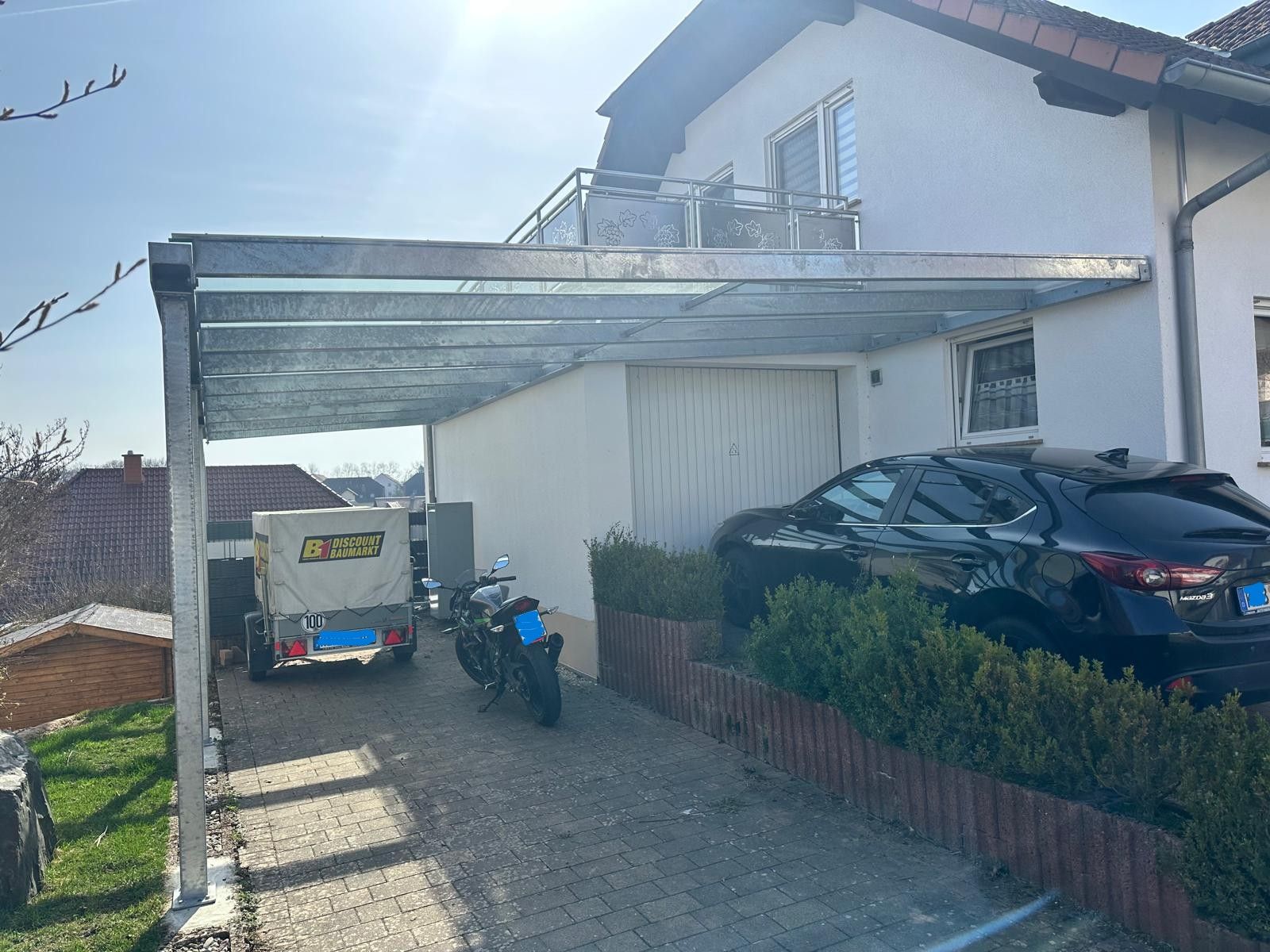 Auffahrt neben einem weißen Haus mit Carport, geparktem Auto, Roller und einem kleinen Anhänger auf einem abfallenden Weg.