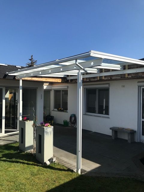 Terrasse mit weißem Haus, Glasdachmarkise und zwei kleinen Pflanzkästen vor den Fenstern