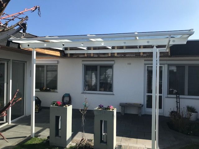 Terrasse mit weißer Pergola, Topfblumen und zwei Podestpflanzgefäßen vor einem Haus