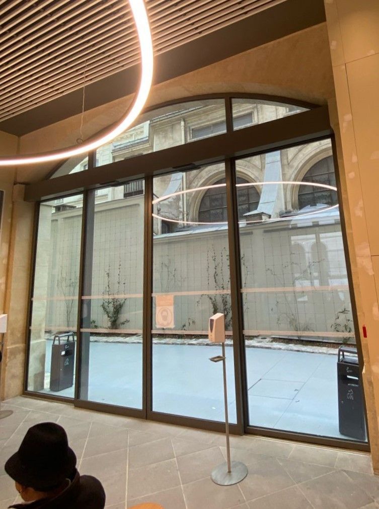 Vue intérieure à travers de grandes portes vitrées automatiques donnant sur une cour intérieure bordée de murs en pierre et sur l'extérieur du bâtiment.