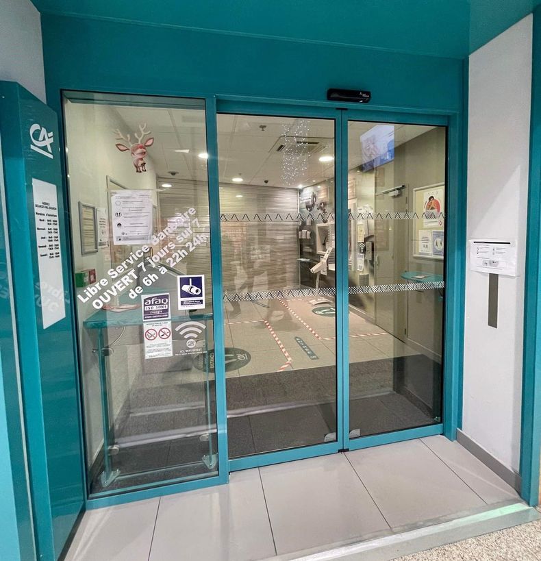 Entrée vitrée d'une agence bancaire Crédit agricole avec encadrement turquoise et panneaux indiquant un accès libre-service 24h/24 et 7j/7.
