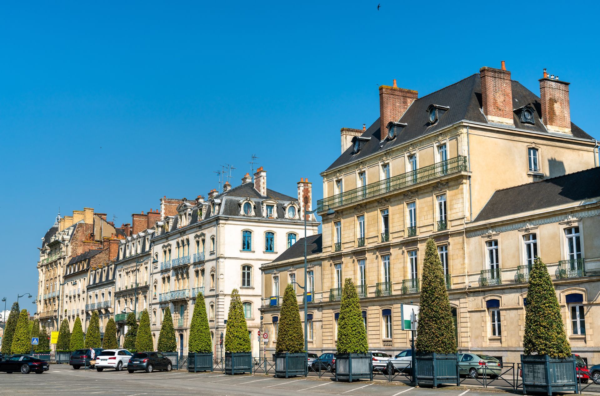 Une rangée de bâtiments historiques aux teintes claires et aux toits gris, bordée de grands conifères en pot, sous un ciel d'un bleu limpide.