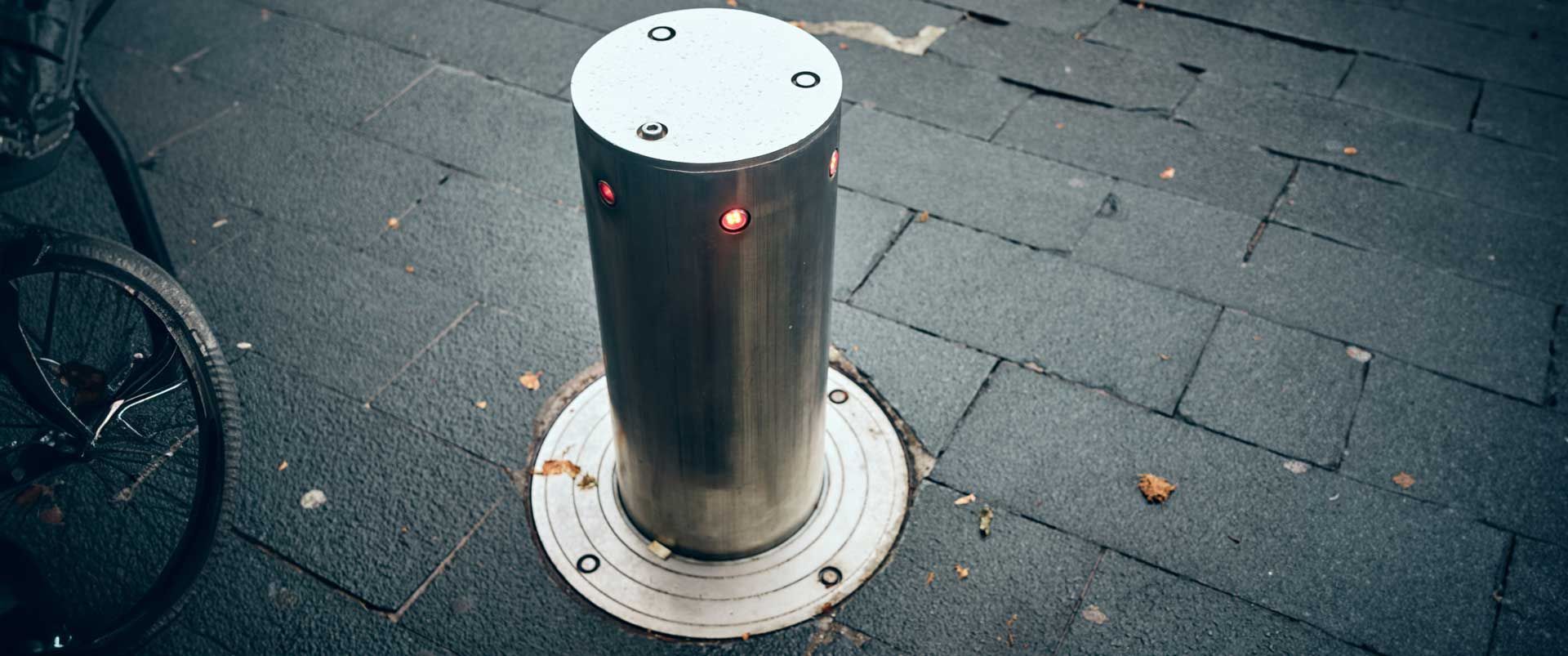 Une borne de sécurité cylindrique en métal se dresse sur un sol pavé, munie de petits voyants rouges lumineux.