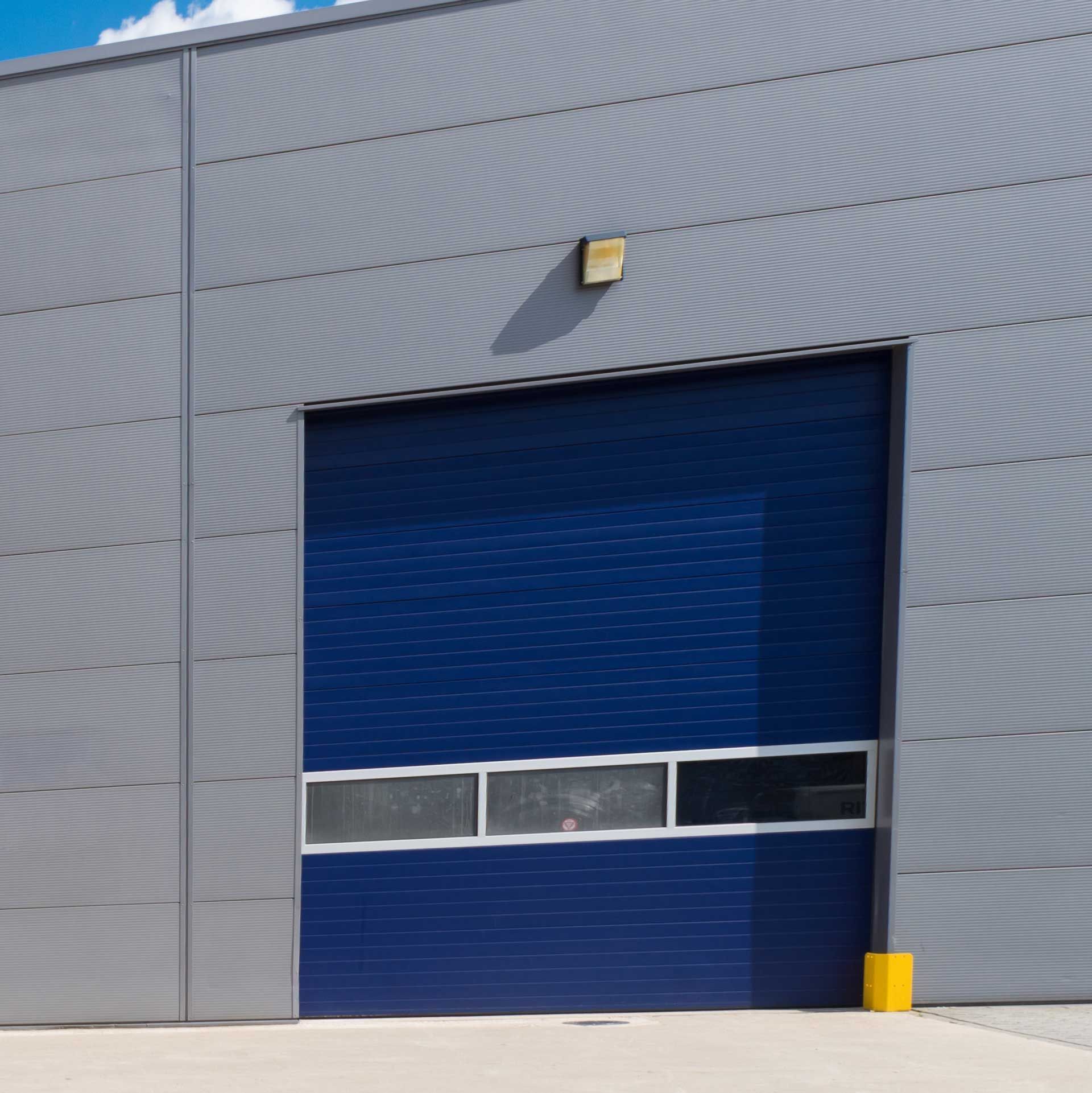 Une porte de garage industrielle bleue avec une rangée de trois fenêtres rectangulaires, intégrée à un bâtiment à façade métallique grise.