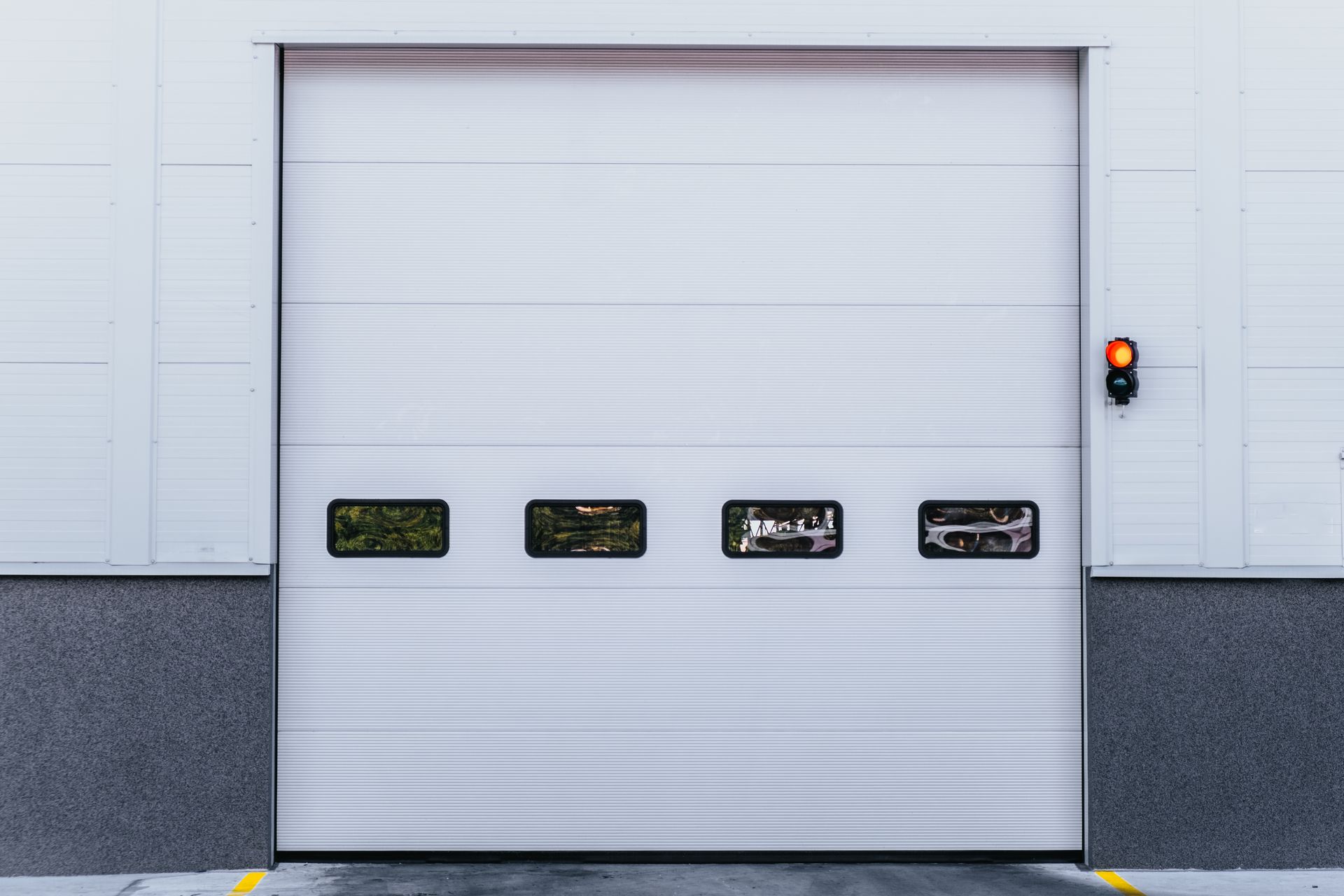 Une porte de garage sectionnelle industrielle blanche fermée, avec quatre petites fenêtres et un feu de signalisation sur le mur de droite.