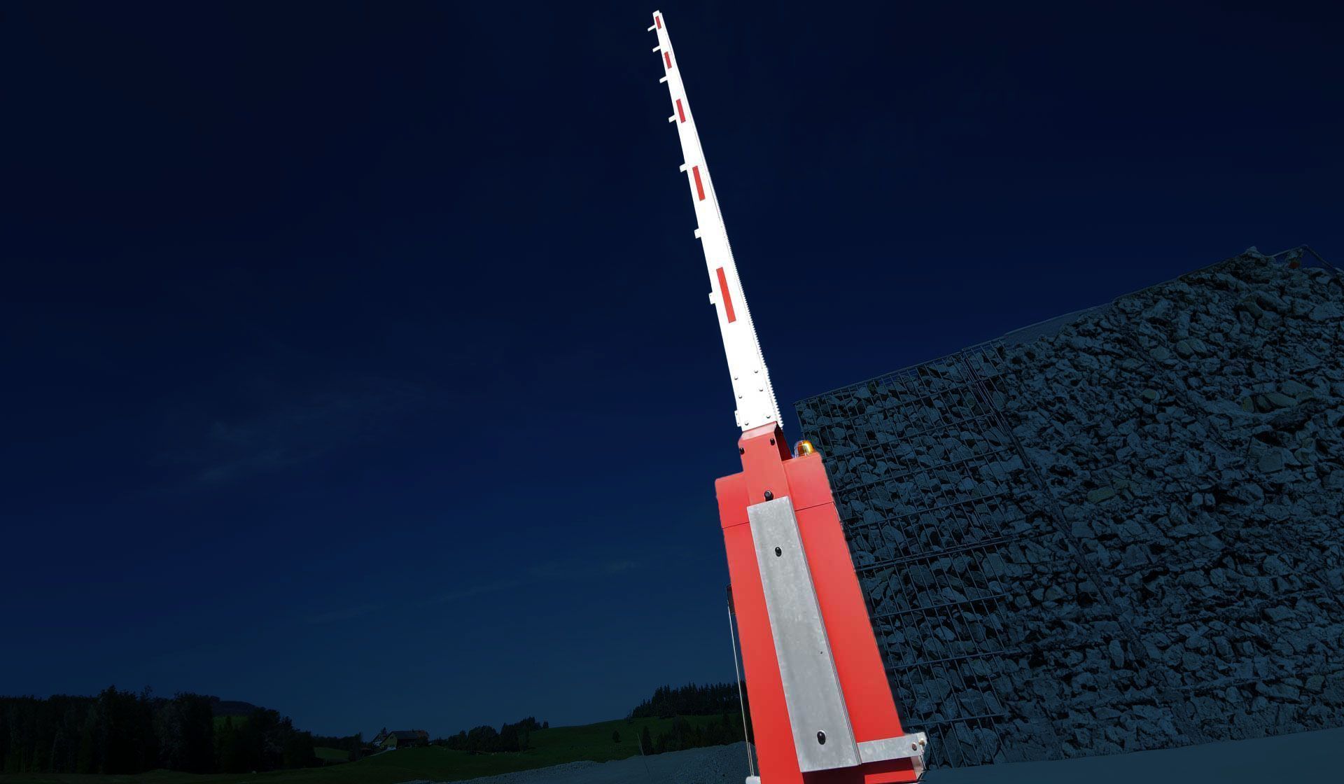 Un bras de barrière industrielle rouge et blanc pointant vers le ciel nocturne, à côté d'un tas de matériaux sombres et texturés.