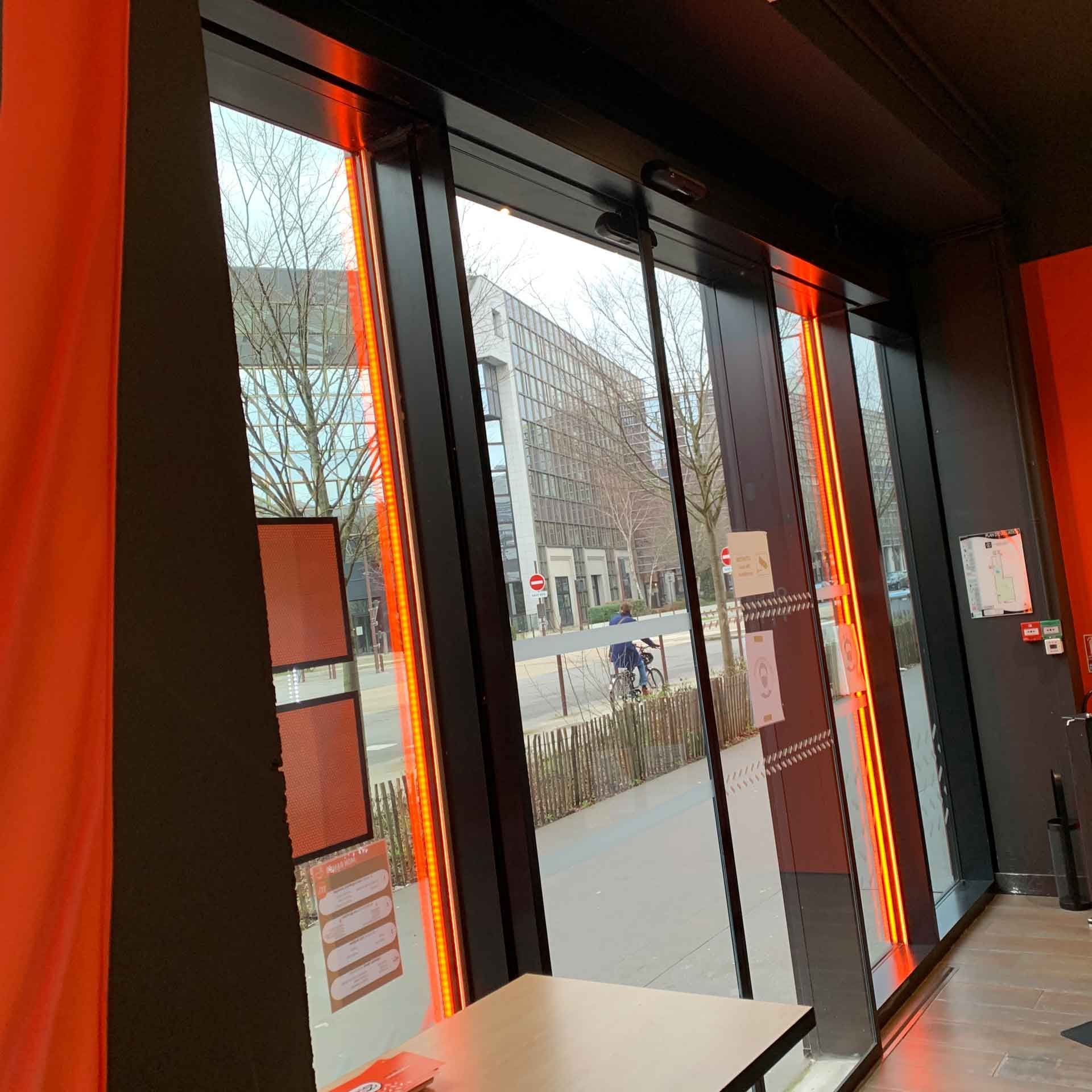 Une vue à travers de grandes portes vitrées, encadrées par des rideaux orange vif, donnant sur une rue bordée d'arbres et de bâtiments.