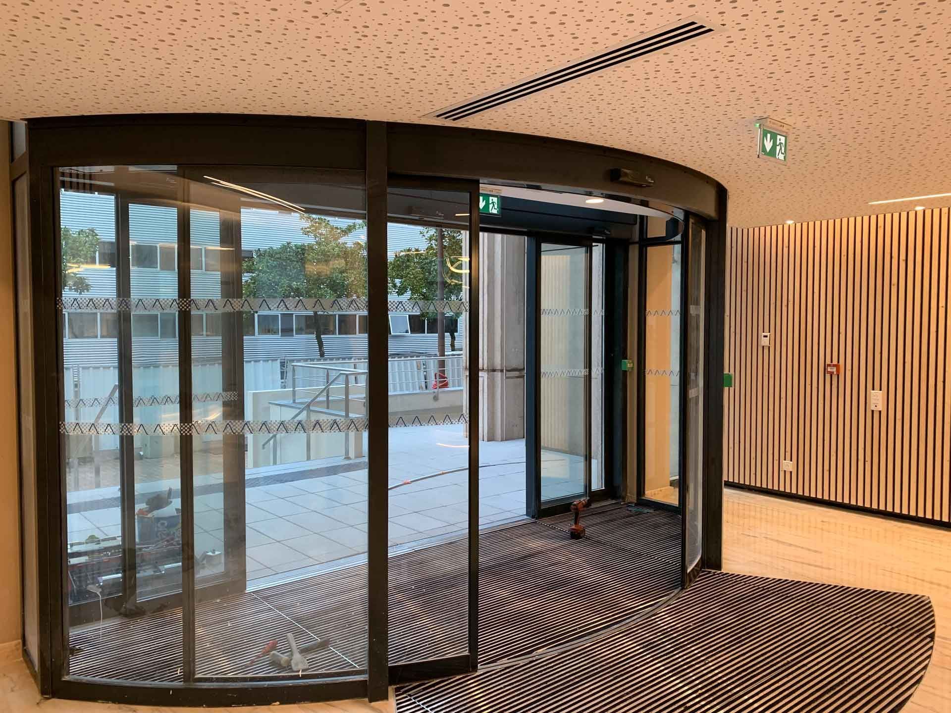Porte tournante en verre incurvé dans le hall d'un immeuble moderne, avec mur à lattes de bois et tapis de sol rayé.
