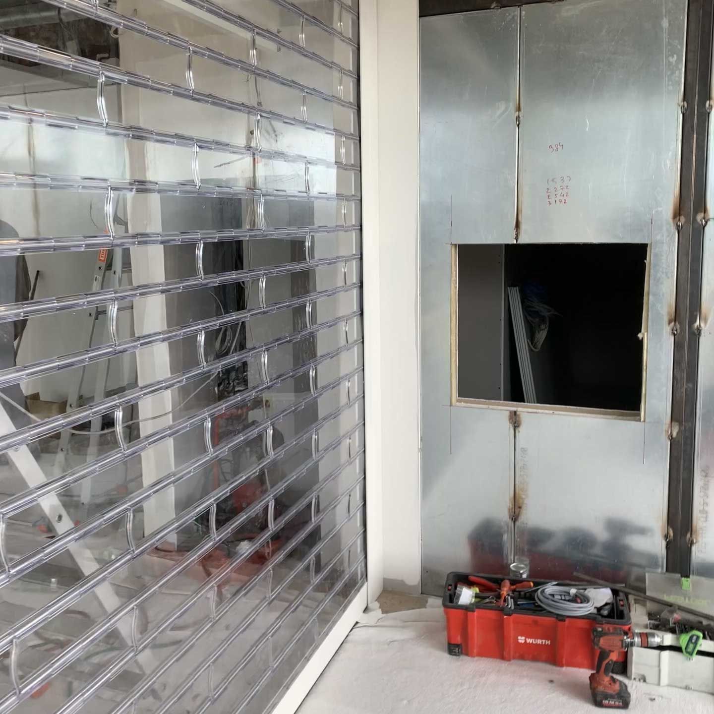 Un chantier de construction avec une grille de sécurité transparente et un mur métallique comportant une découpe rectangulaire, ainsi qu'une caisse à outils rouge.