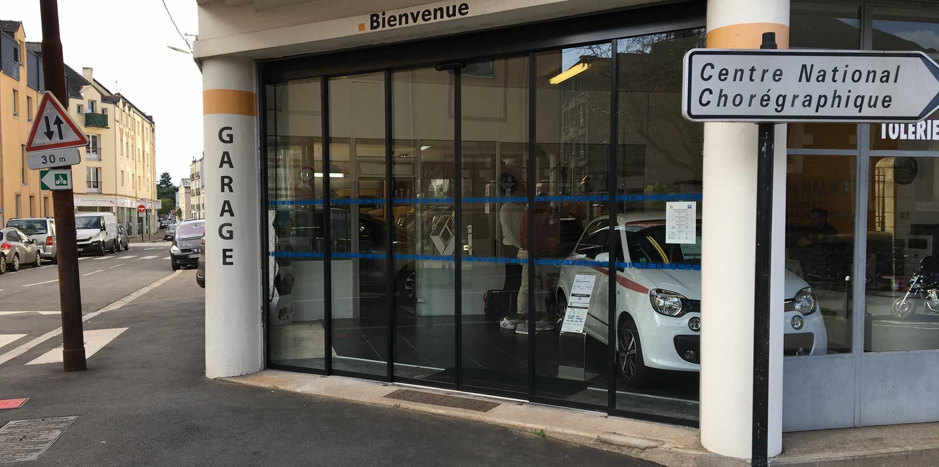 Vue de rue d'un garage à façade vitrée avec une voiture blanche à l'intérieur, à côté d'un panneau indiquant le Centre national de chorégraphie.