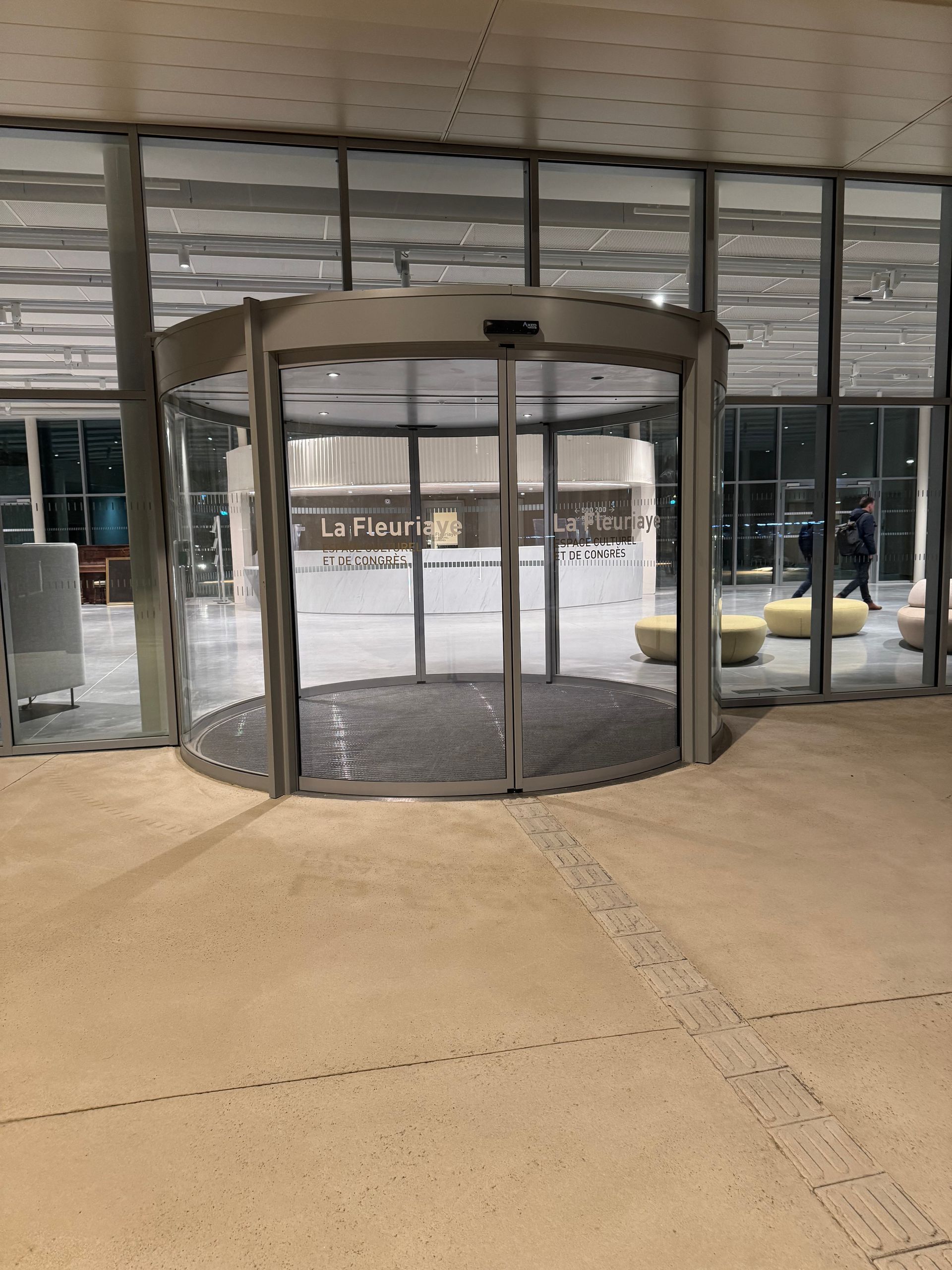 Une porte d'entrée moderne en verre tournant, avec un cadre en métal foncé et un tapis de sol gris devant l'immeuble.