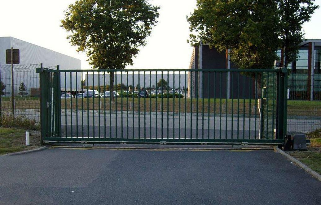 Un portail coulissant en métal vert, installé sur une allée pavée devant des bâtiments industriels et des arbres.