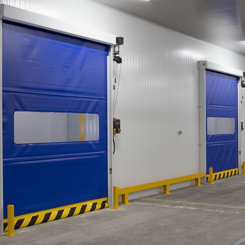 Deux portes roulantes industrielles bleues à grande vitesse, dotées de panneaux de vision transparents, sont montées sur un mur blanc avec des protections de sécurité jaunes.