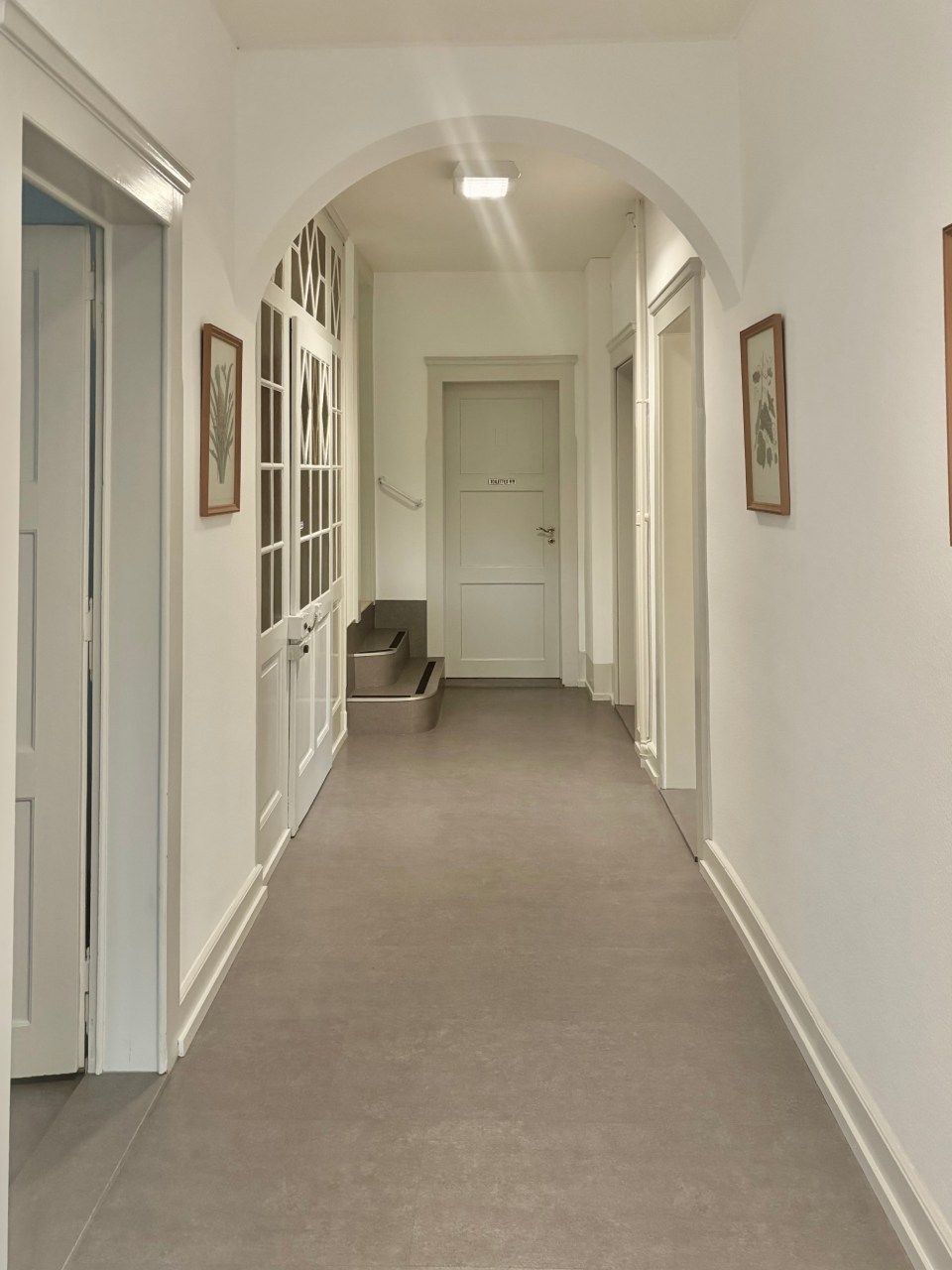 Couloir étroit et blanc avec moquette, plafond voûté, portes et un banc au fond.