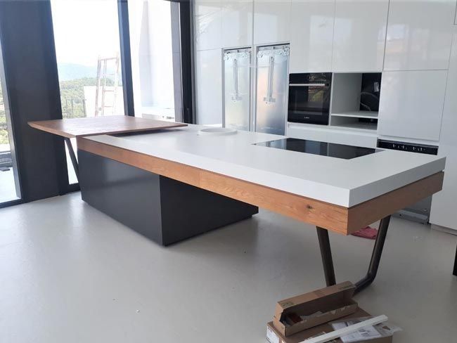 Cocina moderna con una gran isla blanca y de madera, base negra y ventanas luminosas con vista a la ciudad.