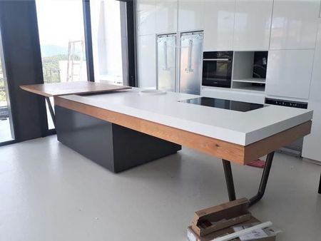 Cocina moderna con una gran isla blanca y de madera, base negra y ventanas luminosas con vista a la ciudad.