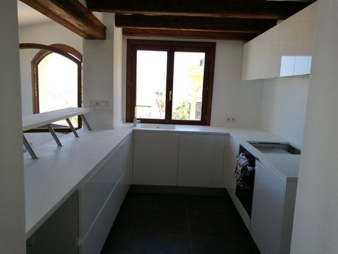 Cocina moderna blanca con suelo oscuro, vigas de madera y una ventana sobre la encimera.