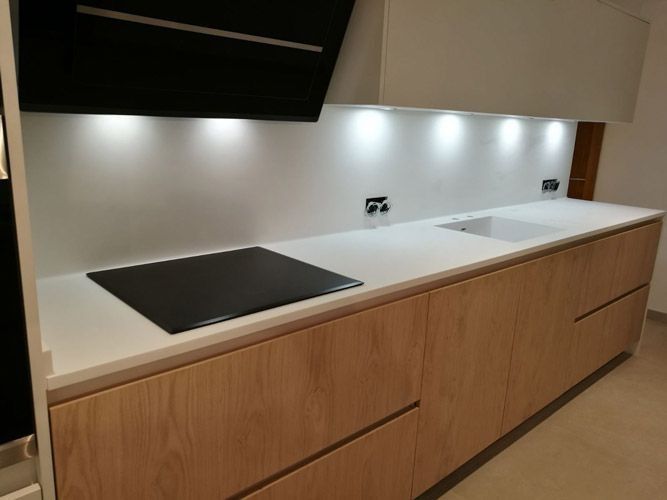 Encimera de cocina moderna con gabinetes de madera, placa de cocción negra y protector contra salpicaduras blanco bajo luces debajo de los gabinetes.