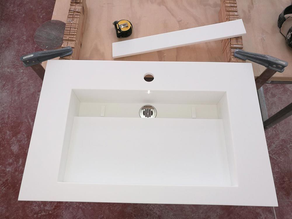 Lavabo blanco con orificio para grifo y desagüe cromado, mostrado sobre una encimera con una cinta métrica y piezas de una lámpara cerca.