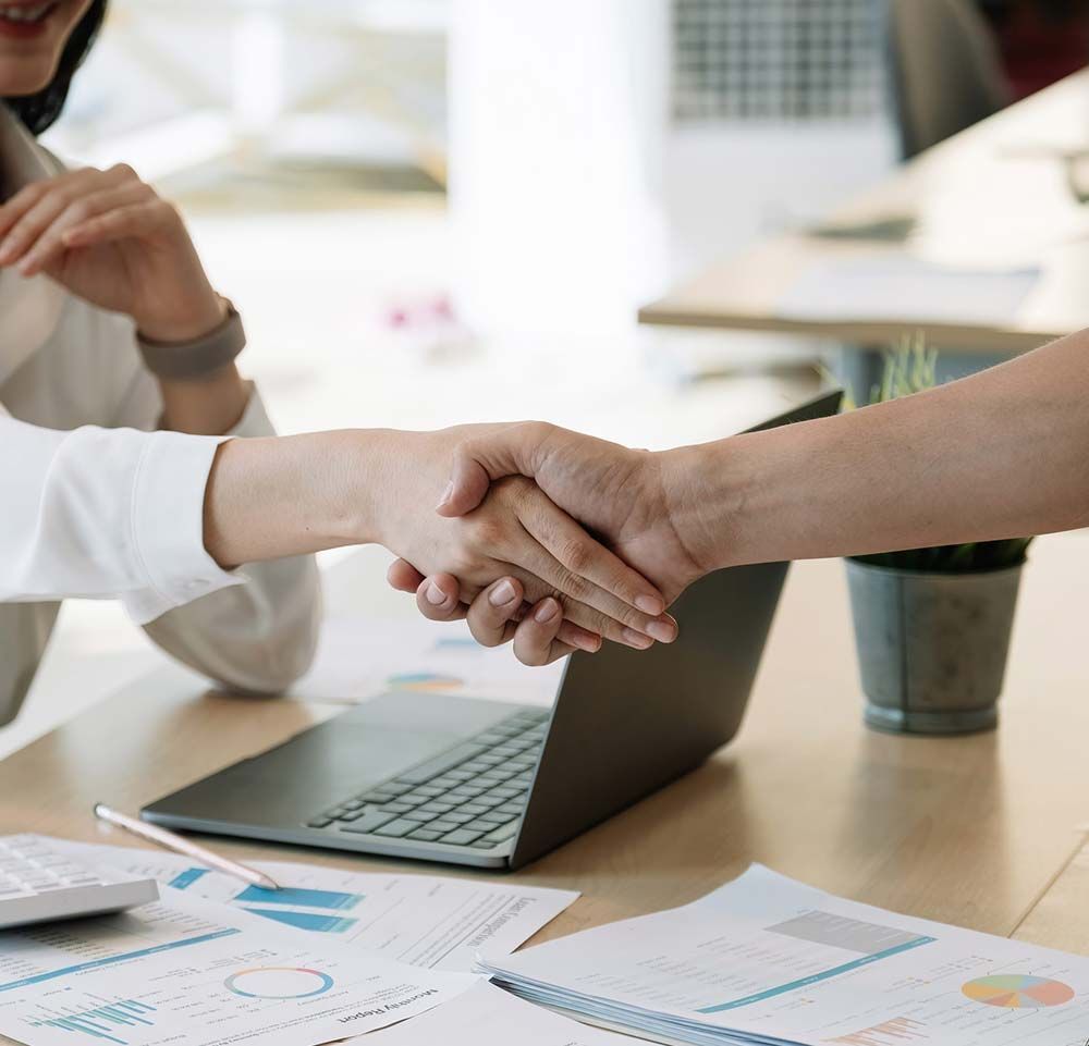 Dos personas se dan la mano sobre una mesa de reuniones con un portátil y gráficos.