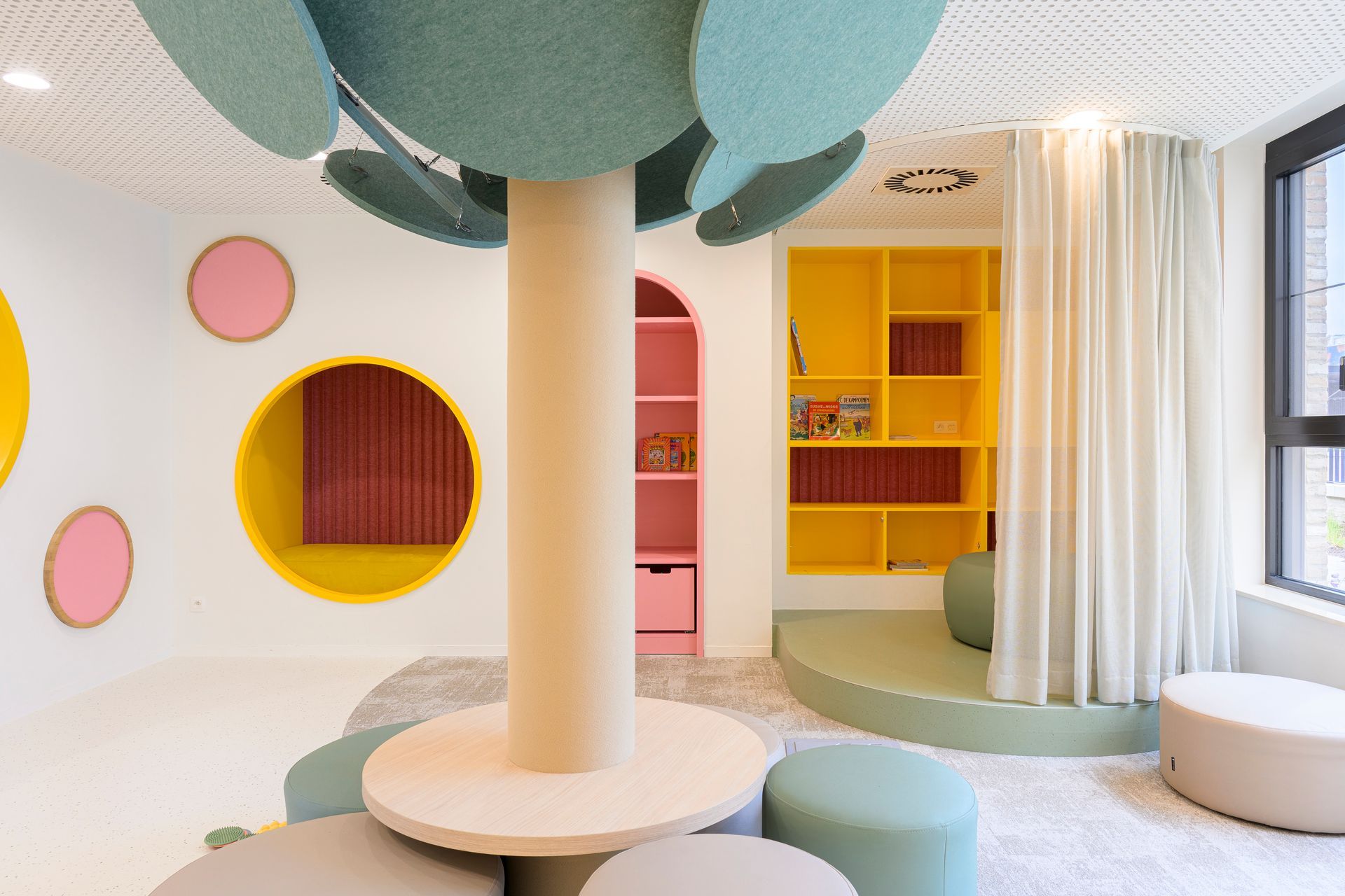 Kleurrijke speelkamer voor kinderen met pastelkleurige ronde uitsparingen, planken en zitkussens rond een centrale pilaar