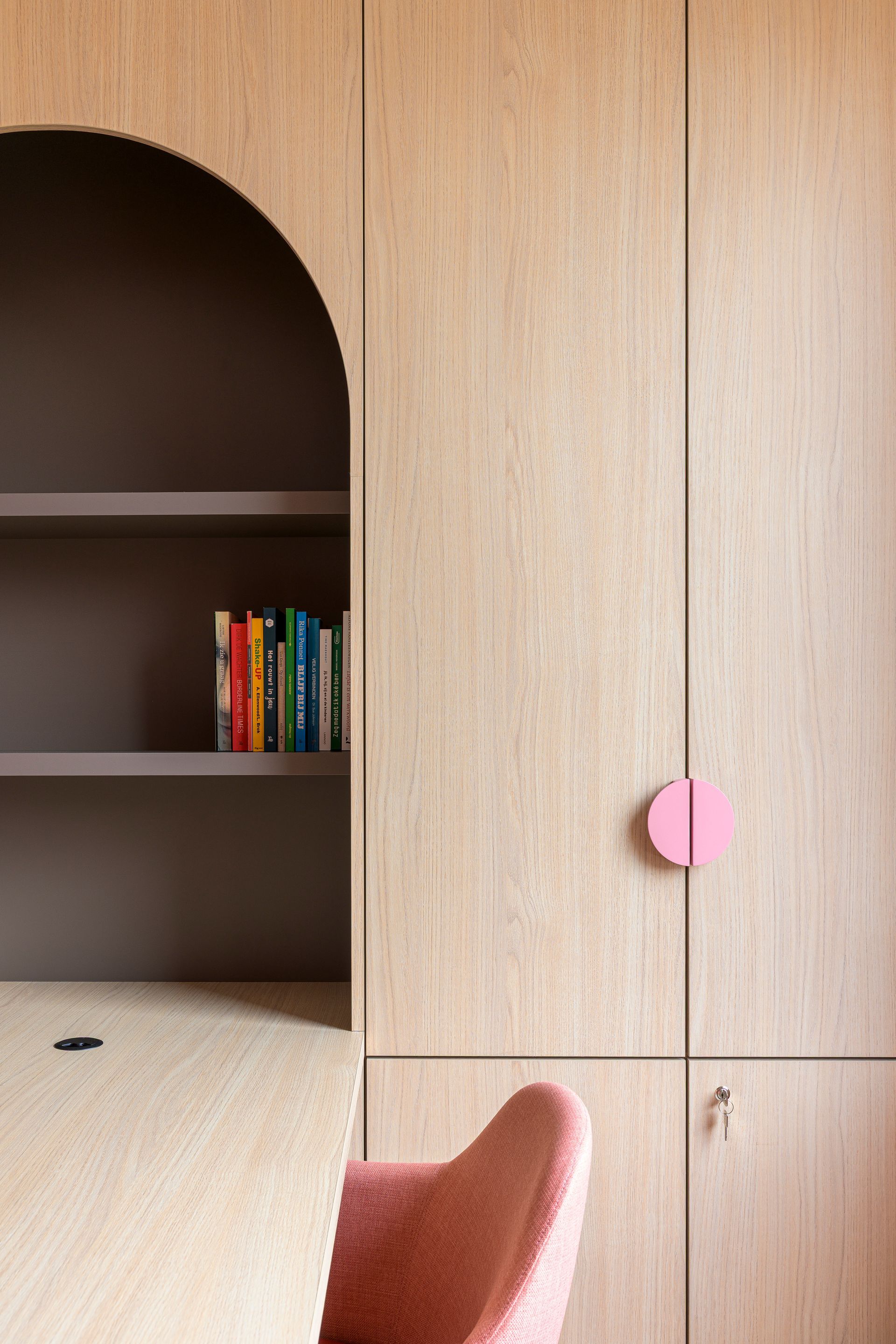Ingebouwde houten kledingkast met roze knoppen naast een gebogen plank en een roze stoel in een minimalistische kamer