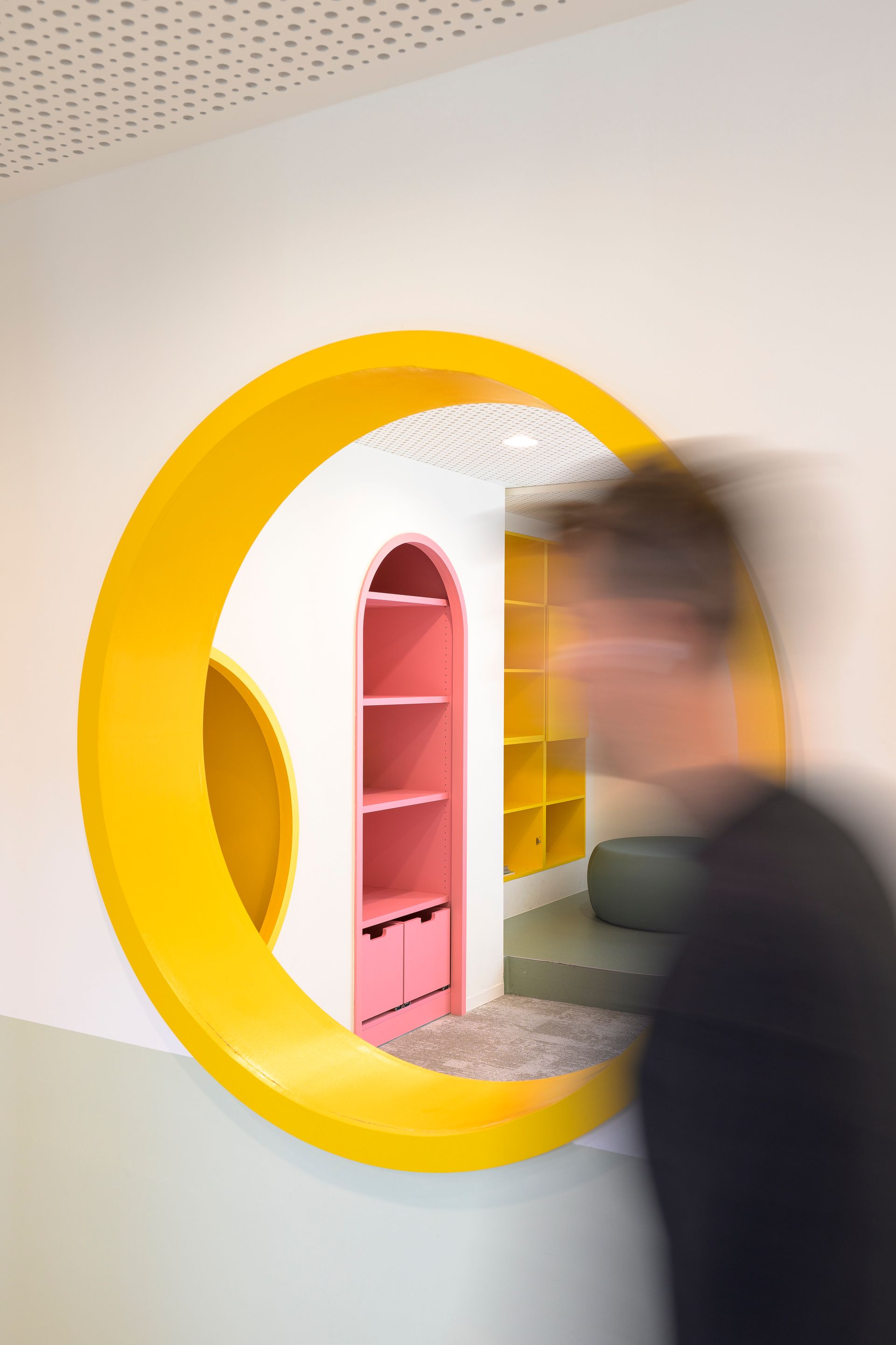 Persoon die langs een felgele ronde opening in de muur loopt, met roze en gele planken erin
