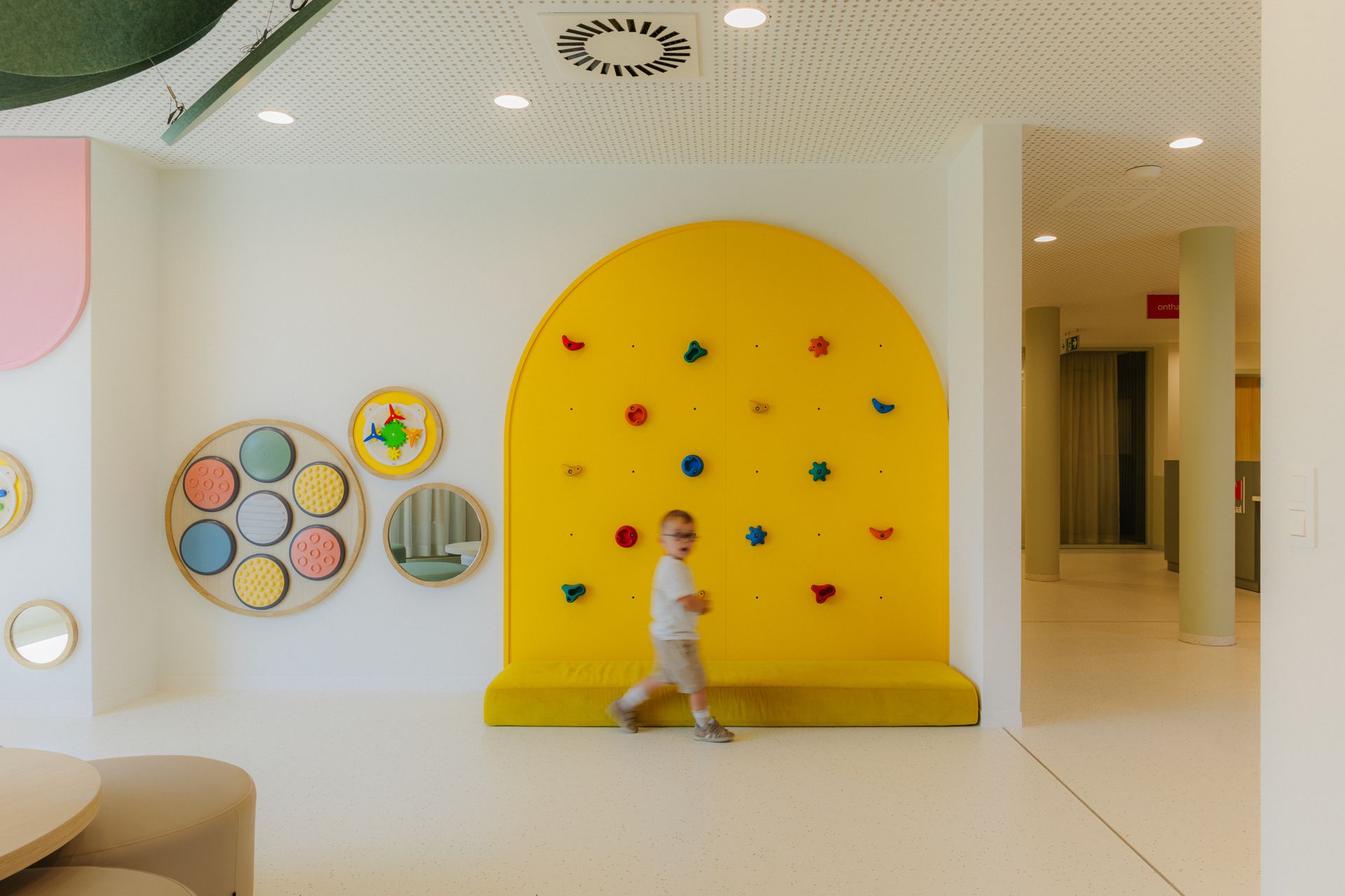 Kind in een lichte speelruimte met een grote gele klimwand en kleurrijke ronde wanddecoraties