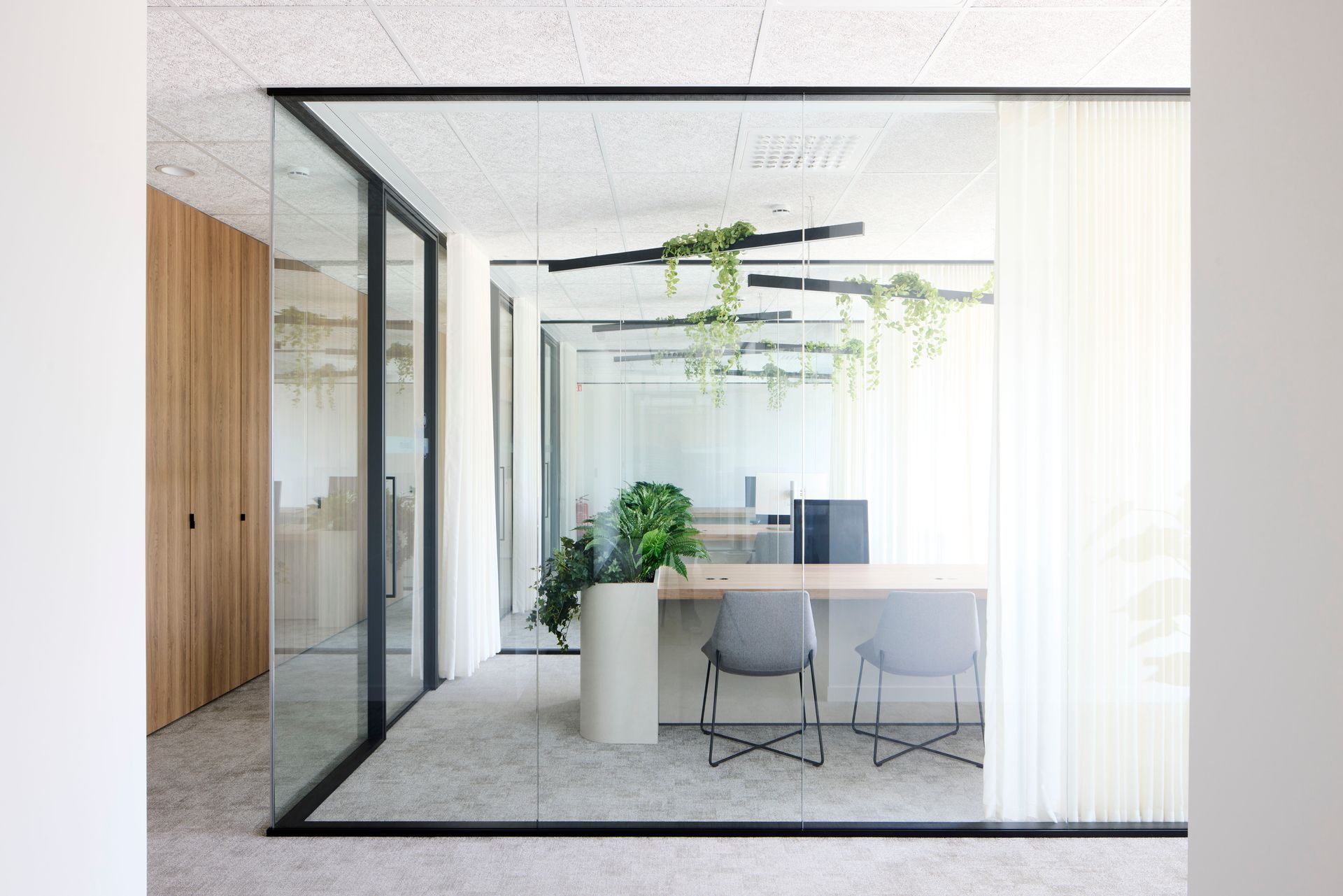 Modern kantoor met glazen wanden, een wit bureau, stoel, planten en hangende groen in een lichte, minimalistische ruimte.