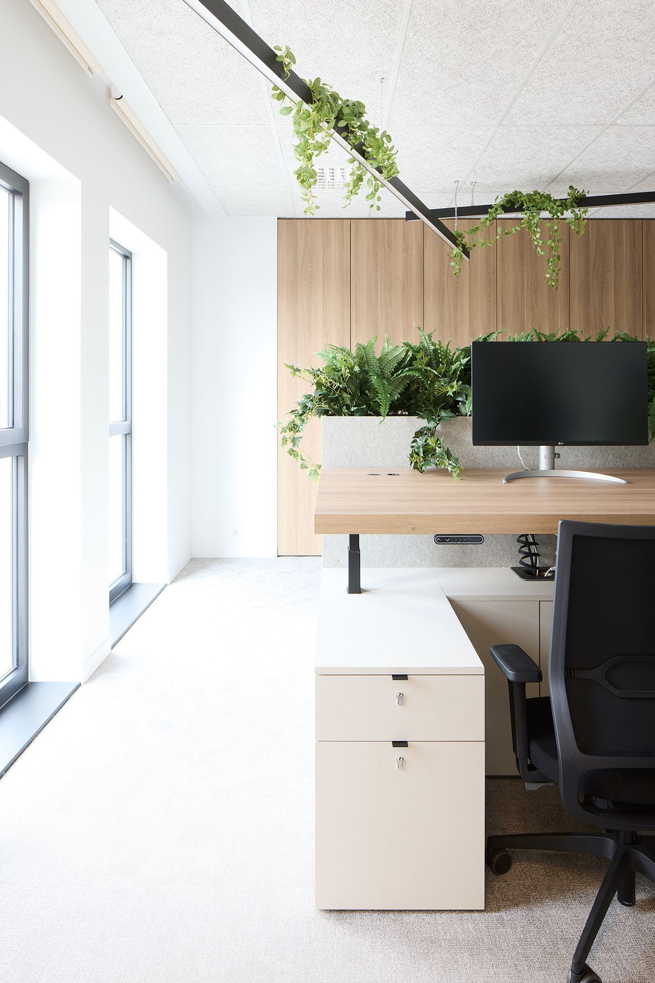 Lichte, minimalistische kantoorruimte met grote ramen, een wit bureau, een zwarte stoel, planten en een aan de muur bevestigde monitor
