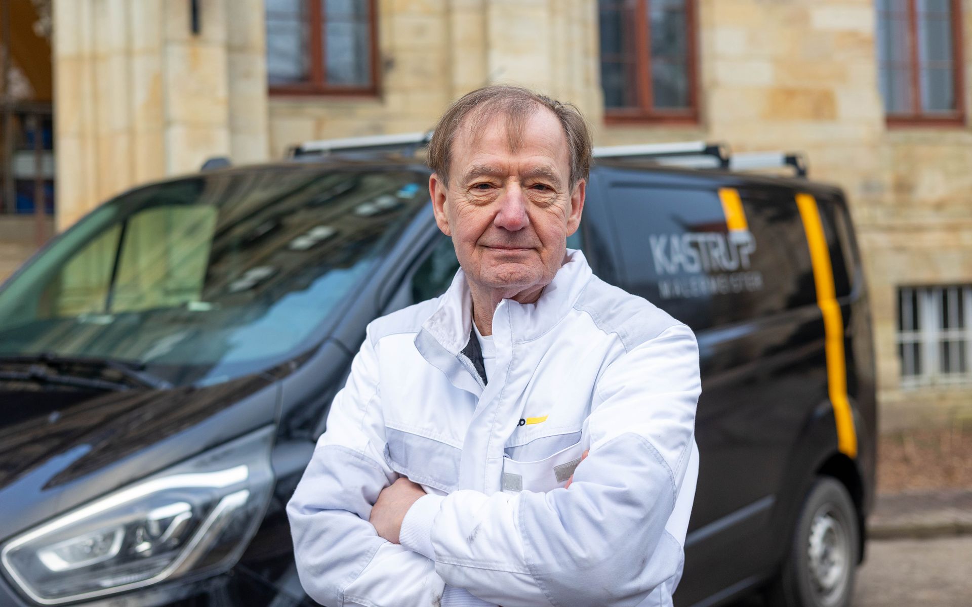 Ein Mann im weißen Kittel steht vor einem schwarzen Lieferwagen.